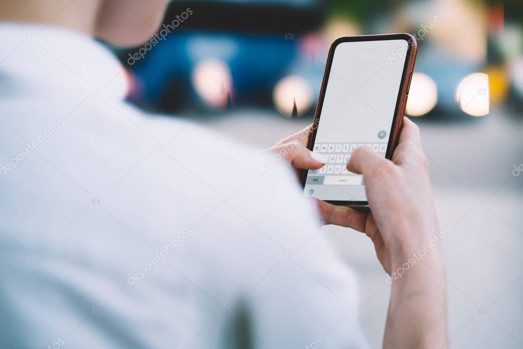 Cropped back view of young woman holding smartphone in hands and typing text on keyboard of modern device with blank display.Hipster blogger messaging with followers on telephone with blank screen