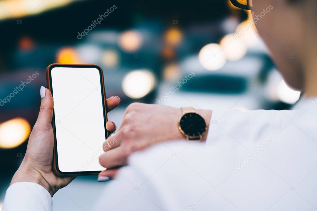 Cropped back view of woman's hand holding smartphone with blank screen for your internet website and checking time on watch on another hand on bokeh background.Digital telephone with copy space area