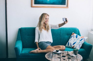 Hippi kız mutlu kadın fotoğraf yolu ile smartphone aygıt ev iç çekmek, sehpa, önünde rahat kanepe otururken telefonla örümcek ağı fotoğraf makinesi ile pozitif selfie fotoğraf yapma gülümseyen