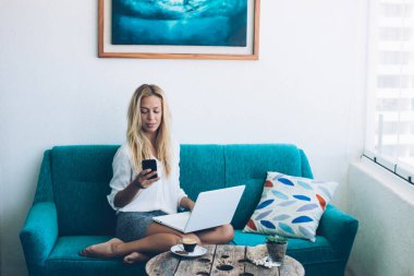 Çekici kadın online ödeme hareketine ev iç rahat kanepede oturan modern smartphone aygıtında teyit ederken dizüstü aygıtta alışveriş sırasında web mağaza öğeleri seçme