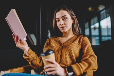 Otodidakt ile edebiyat kahve dükkanında, kafein içecek ve ders kitabı düşman eğitim tutan kameraya arayan ciddi kadın için vakit konsantre zeki kız öğrencinin portresi