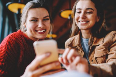 İki gülümseyen kadın blogcular mutlu bin yıllık kadınlar üzerinde boş zaman teknoloji ile dinlenme modern cep telefonu aracılığıyla arkadaşlarınızla sohbet cep telefonu, fotoğraf uygulaması kullanarak yayın oluşturma