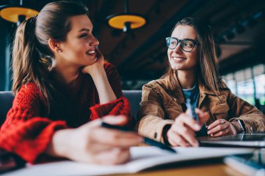 Birbirlerine bakarak ve göz teması kapalı, gülümseyen kadın ev ödevi için araştırma Üniversitesi Kütüphanesi'nde birlikte yapmak öğrenme sırasında yakalamak canlı iletişim zevk olumlu hipster kızlar