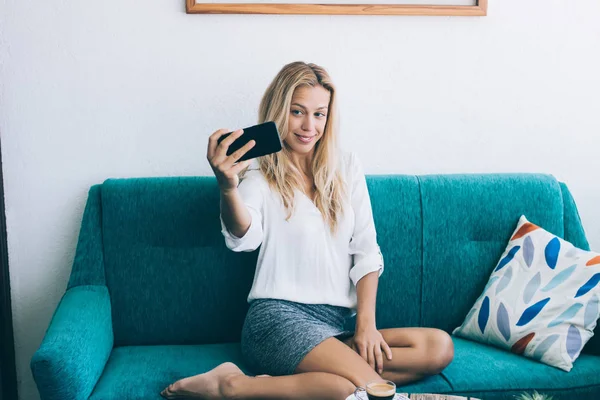 Cheerful hipster girl making positive photos for publication in social networks using mobile phone indoors, successful female blogger taking pictures via media application on smartphone device