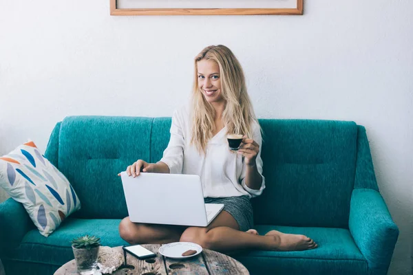微笑的时髦女孩的肖像使用 Wifi 连接和膝上型计算机设备在家庭内部 当看照相机和拿着杯子与咖啡 开朗的妇女更新软件在现代上网本 — 图库照片