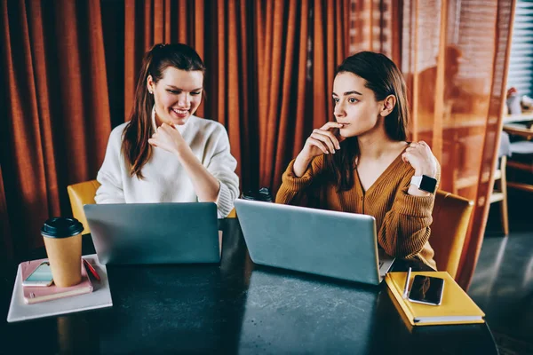 两位成功的女程序员坐在咖啡馆 享受着与上网本的距离工作 积极的千禧时髦女孩使用 Wifi 在笔记本电脑上搜索信息 — 图库照片