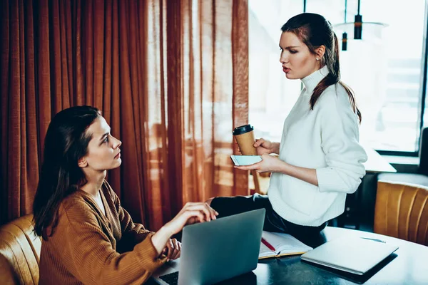 Millennial female freelancers discussing ideas for teamwork during coffee break at cafeteria, young serious woman holding smartphone and explaining to friend how checking updates on laptop computer