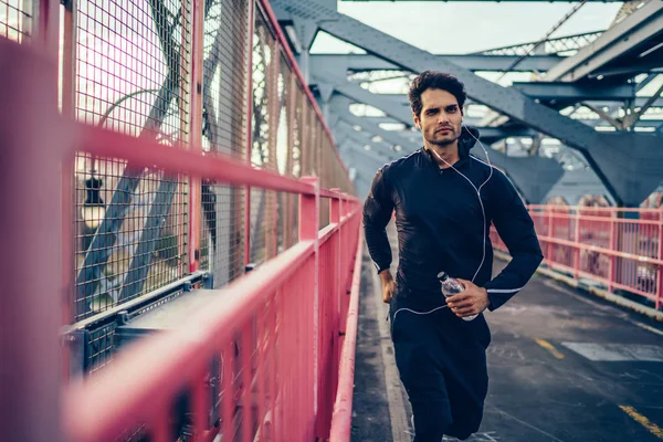 Young man in sportswear jogging on city bridge with bottle of water in hands, european athlete running on urban setting and listening playlist via earphones, active health and motivation lifestyle