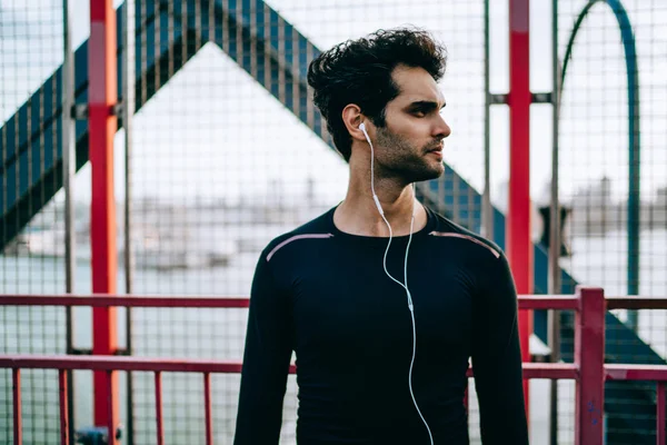 Handsome male runner in electronic earphones looking aside after healthy training on urban setting, concentrated man in sportswear listening serious audio record about cardio exercises via headphones