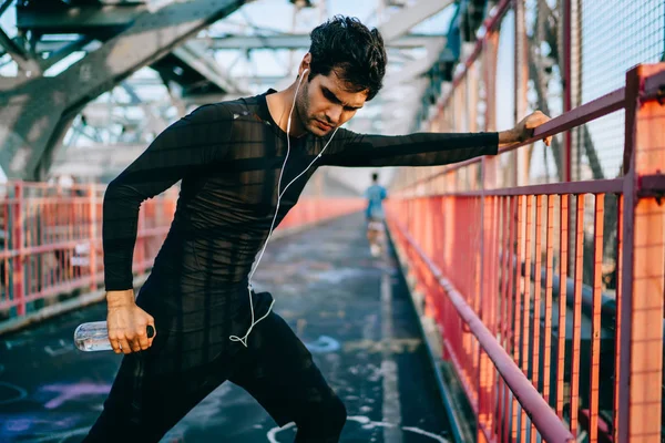 European male jogger in headphones doing stretch exercises for all body muscles before start training outdoors, young flexible man in sportswear holding bottle with water and workout on city bridge