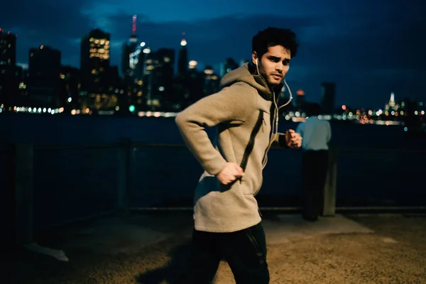 Male runner in earphones doing cardio exercises for making heart rate up and looking at camera during workout process, young sportsman jogging outdoors for strengthen body muscles and improve mobility