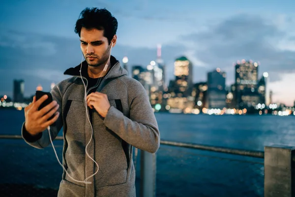 Concentrated male runner making high volume via smartphone device for listening training playlist via earphones before jogging outdoors, serious sportsman standing with cellular on blurred background