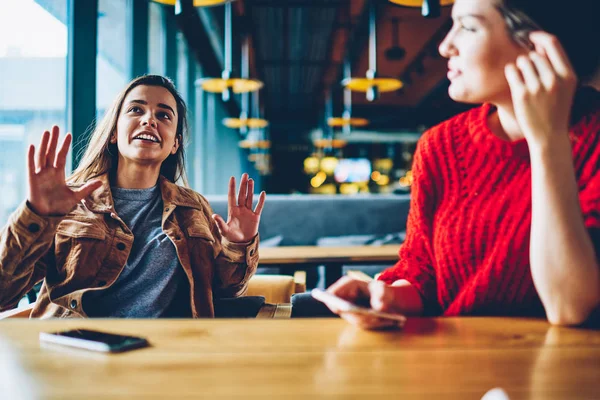 Two happy hipster girls discussing ideas for attracting followers for shared blog in social networks indoors, successful female users communicated at comfortable cafeteria while waiting friend