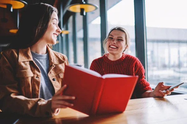 Two excited female bloggers discussing positive best seller sitting at table in cafeteria, successful hipster girls reading interesting literature enjoying free time on leisure at university campus