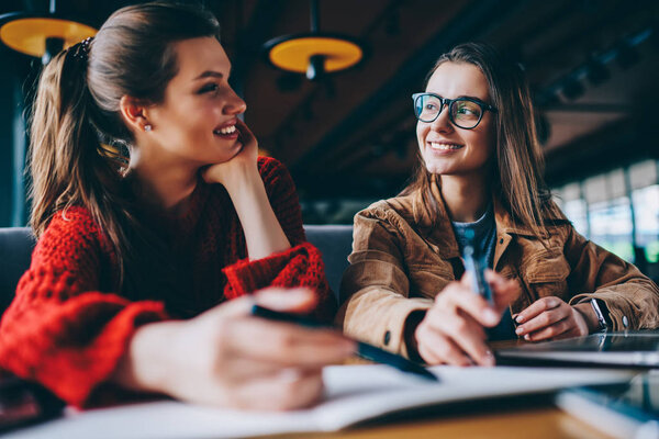 Positive hipster girls enjoying live communication looking to each other and catching eye contact during learning indoors, smiling women making research for homework together at university library