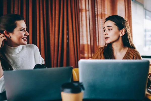 Millennial female friends making positive conversation in break of working on netbooks resting in wifi zone with coffee beverage, successful hipster girls recalls to pleasant moments spending together