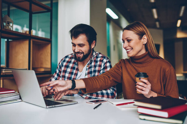 Cheerful internet users laughing and enjoying time after watching webinar online at coworking space, smiling friends sitting at cafeteria and communicated via laptop while girl holding cup coffee