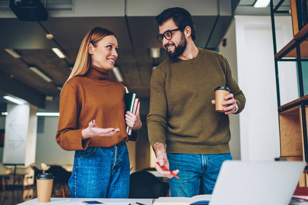 Two business coworkers 20 years old communicated positively in comfortable office during coffee break, handsome hipster guy in spectacles holding drink while attractive girl staying with books indoors