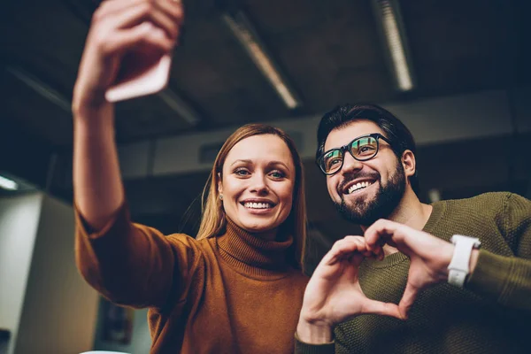 Happy bloggers makes funny picture for profile to attract new followers to social networks accounts while girl clicking photo  of themself during bearded guy in glasses posing and shoving \'heart\'