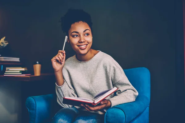 Smiling creative female blogger pondering on idea for publication sitting in comfortable armchair,young african american woman recalling information while planning to do list at home interior