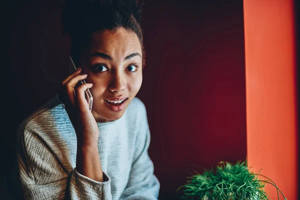 Portrait of emotional dark skinned woman shocked with information get during mobile phone conversation, young african american female expressing surprise looking at camera talking on cellula
