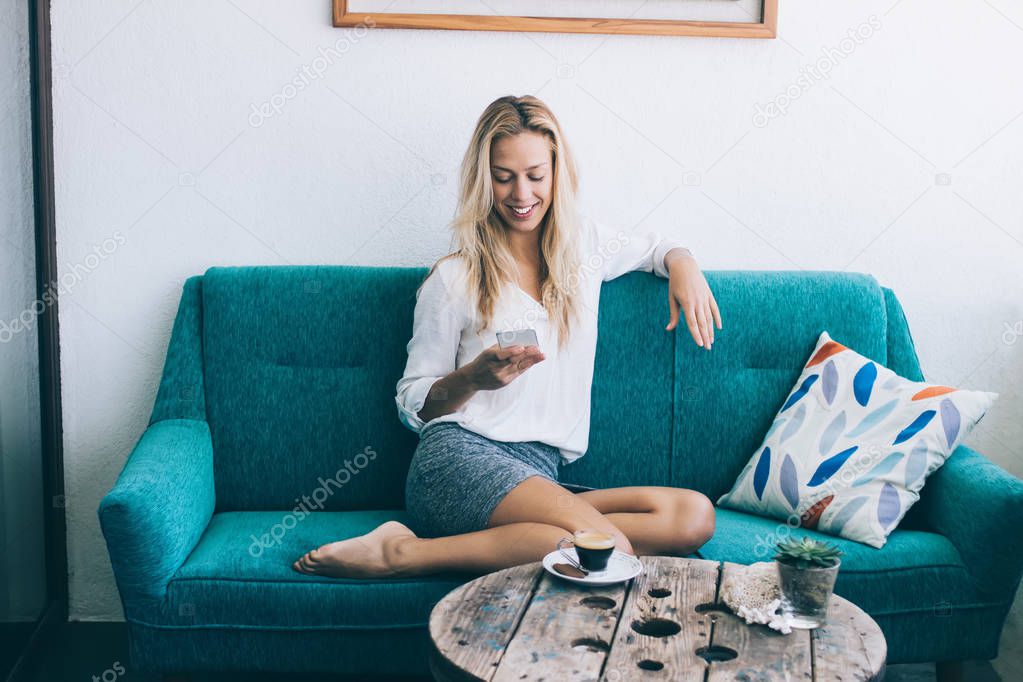 Cheerful female blogger creating publication for social networks via multimedia application on smartphone device while sitting at cozy couch with comfort, smiling woman reading friendly email
