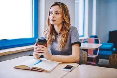 Üniversite için makale üzerinde okul sırası vasıl defter ile oturma ve eğitim hakkında düşünme paket servisi olan restoran Kupası tutan genç dalgın kadın kapalı, kahve molası sırasında durulması düşünceli kız öğrenci