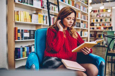 Çekici beyaz hippi kız kitapçı, sandalyede oturan edebiyat hobi için vakit konsantre sevimli kız öğrencinin Üniversite Kütüphanesi'nde ilginç en çok satan okuma zevk