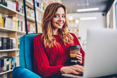 Portrait of positive hipster girl looking at camera holding takeaway cup with coffee and enjoying studying on laptop device at bookstore, cheerful female student sitting at library and using computer
