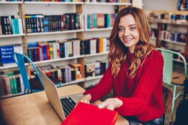 Cheerful hipster girl taking notebook for education sitting at desktop with modern laptop device, positive smiling female student spending time at modern bookstore for making research for project clipart