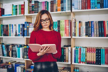 Clever female journalist searching information for article standing with literature with bookshelves on background, attractive young woman in eyeglasses reading best seller in college university clipart