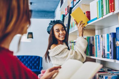 Kameraya bakarak ve edebiyat raflardan portre pozitif kadın arkadaş ile vakit için arama en iyi satıcı seçme yüzünde sevimli gülümseme ile genç başarılı Afrika hippi kız