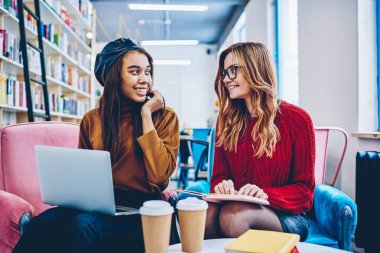 Modern dizüstü bilgisayar ve eğitim için ders kitabı ile çok kültürlü başarılı hipster kızlar kapalı e birlikte öğrenme sırasında iletişim Üniversitesi kütüphanesinde oturan gülümseyen kız öğrenci