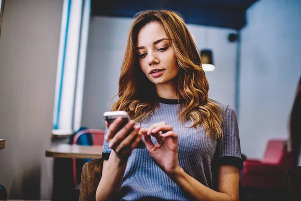 Concentrated female user reading serious text message via application on modern cellular phone, attractive hipster girl typing email during communicate with followers in social networks via smartphone