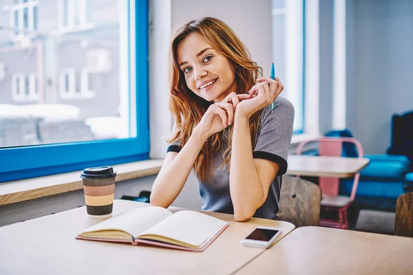 一个开朗的时髦女孩的肖像看着相机 而坐在木制桌面与外卖咖啡和教科书的教育 快乐微笑的女人享受天生在大学校园 — 图库照片