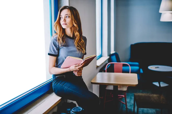 Dreaming hipster girl spending time at campus for literature hobby pondering on romantic best seller, thoughtful calm female teenager holding textbook for education sitting on cafeteria windowhill