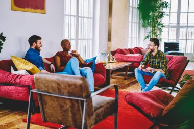 Neşeli grup çok ırklı arkadaşıyla eğlenmek serbest zaman sırasında modern loft iç daire, düz tartışırken kırmızı mobilya oturup konuşmayı hipster çocuklar hafta sonu için plan-