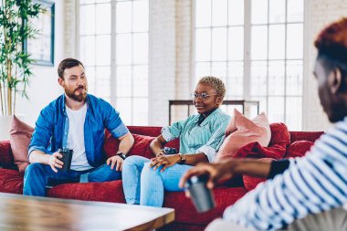Sakallı adam ile kahve fincanı çatı iç coworking alanı, çeşitlilik mürettebat birlikte dinlenme ve boş zaman couc üzerinde oturan üzerinde fikirleri tartışmak meslektaşlarının konuşma sırasında arkadaş arıyorum