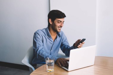 Ofiste oturan ve mutlu adam bilgi dizüstü bilgisayarda arama sonra smartphone, bildirim kontrol çatırdamak ile sokulgan yolu ile hücresel bağlantı, işin sonu zevk olumlu iş adamı