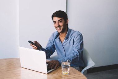 Gülümseyen başarılı erkek grafik tasarımcı vasıl okul sırası ofiste bekleyen webinar sırasında kamera bakıyor ve akıllı telefon elinde tutan olumlu adam dizüstü aygıtı modern çalışma alanında çalışma