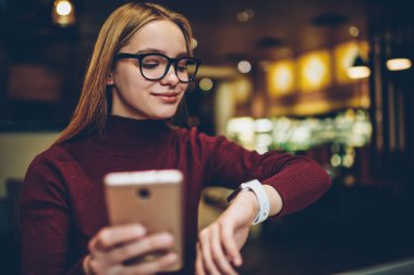 Giyilebilir elektronik kol saati sırasında bekleyen arkadaş kafede zamanında kontrol ve cep telefonu aracılığıyla, smartwatch ekrana bakarak gözlük başarılı hippi kız tebliğ pozitif kadın