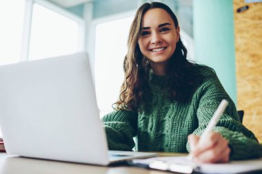 Netbook ile ödev gülümseyen kız öğrenci portresi online ders ile Not Defteri'nde yazma ve araştırma dizüstü bilgisayarda yapmaktan kamera bakarak neşeli esmer kadını tatmin.