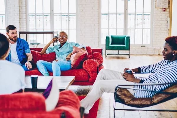 Smiling multiracial group of friends sitting on modern sofa recreating and talking to each other at apartments ,positive male and female hipsters communicating in lounge zone of living room