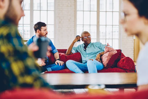 有选择地专注于坐在沙发上喝咖啡杯的非洲裔美国人当她的朋友们交谈时 多种族的同事们在现代设计的 Coworking 空间休息休息 — 图库照片