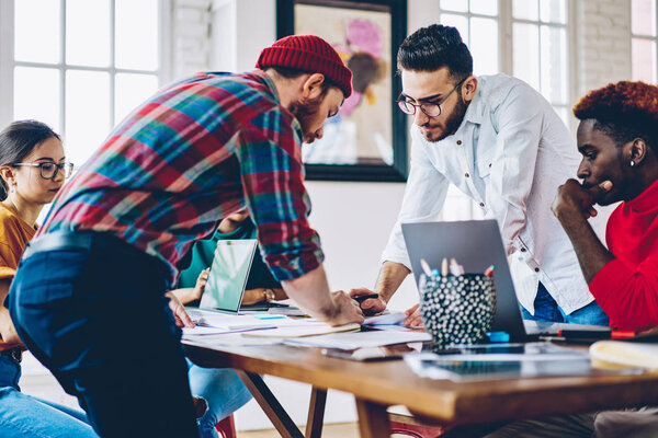 Серьезные коллеги-архитекторы мужского пола проверяют эскизы и бухгалтеры, работающие в команде, многорасовая команда сотрудников сосредоточилась на планировании проектов, сидя за рабочим столом и обсуждая идеи
