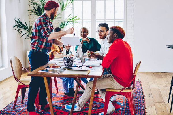 Skilled male employee presenting his strategy to multiracial crew of colleagues sitting together at meeting table, young creative team discussing ideas and opinions cooperating on project planning