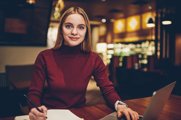 Portrait of positive millennial hipster girl sitting with textbook and laptop computer and preparing to university project, smiling woman looking at camera while writing content article for blog
