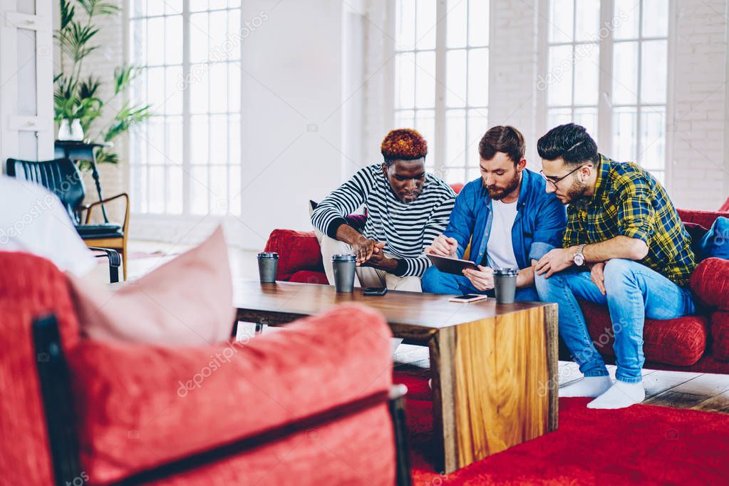 Concentrated multiracial men choosing items in web store during free time together via tablet,  young hipster guys watching movie on portable pc connected to wireless internet in coworking spac
