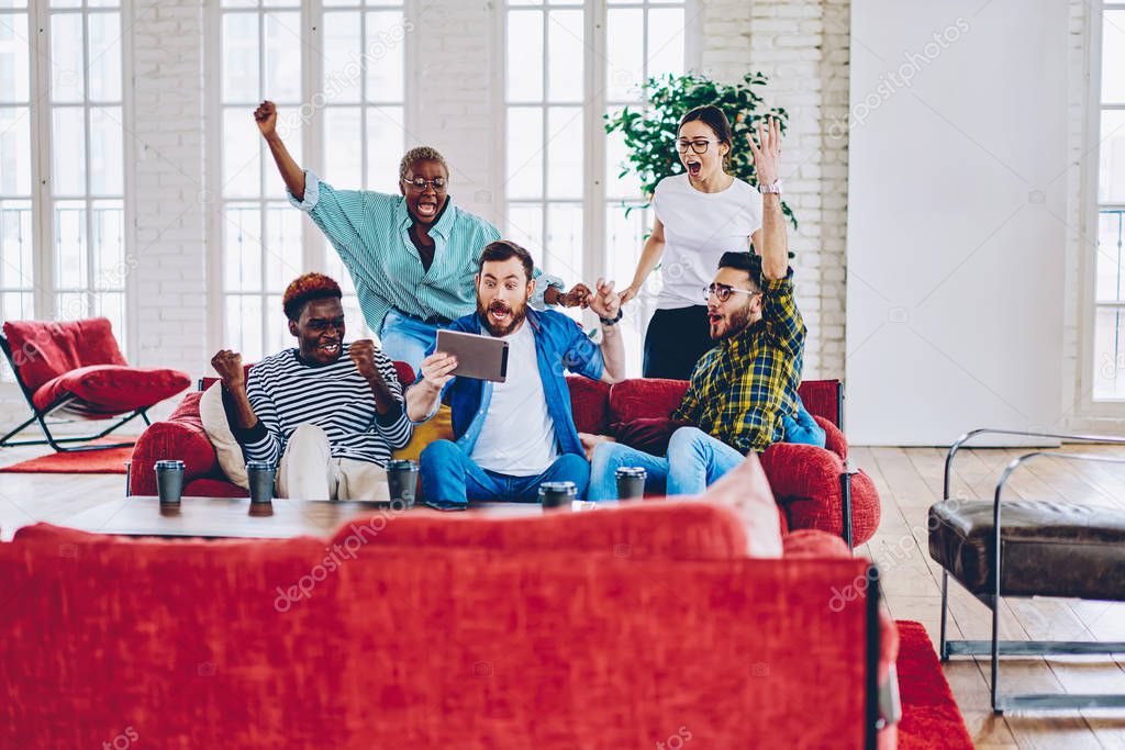 Overjoyed multiracial group of male and female fans cheering for soccer game watching on digital tablet together in loft interior, happy emotional hipsters expressing surprise with victory of contes