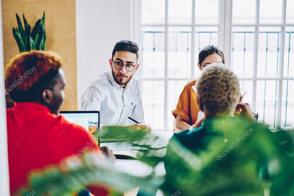 Professional male employee in eyewear looking at colleagues during meeting table discussing cooperation details, diversity group of creative designers sitting on conference about planning projec